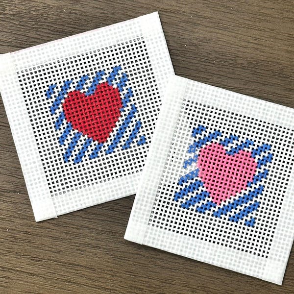 Può includere: Due sottobicchieri quadrati a punto croce. Uno presenta un cuore rosso, l'altro un cuore rosa. Entrambi i cuori sono circondati da strisce diagonali blu su sfondo bianco. I sottobicchieri sono realizzati in un materiale simile a una rete.