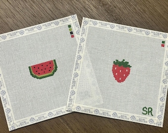 May include: Two square needlepoint canvases with white mesh backgrounds. One canvas features a watermelon slice with a green rind and red flesh with black seeds. The other features a red strawberry with green leaves and the green letters SR.