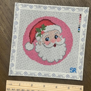 May include: A square needlepoint canvas featuring a circular pink background with a Santa Claus illustration wearing a red hat with holly. The border displays the text SOUTHERN MADE repeatedly. A wooden ruler shows the canvas is 5 inches wide.