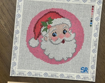 May include: A square needlepoint canvas featuring a circular pink background with a Santa Claus illustration wearing a red hat with holly. The border displays the text SOUTHERN MADE repeatedly. A wooden ruler shows the canvas is 5 inches wide.