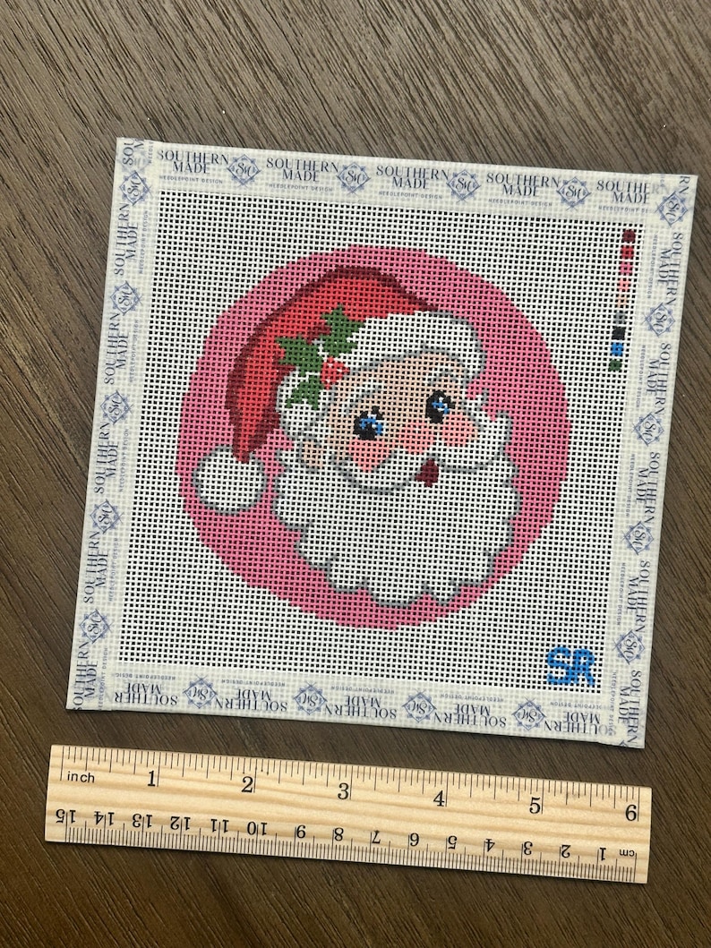 May include: A square needlepoint canvas featuring a circular pink background with a Santa Claus illustration wearing a red hat with holly. The border displays the text SOUTHERN MADE repeatedly. A wooden ruler shows the canvas is 5 inches wide.