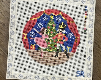 May include: A square needlepoint canvas featuring a circular design of a ballerina in a pink dress and a nutcracker standing before a Christmas tree. The scene is framed by red curtains against a blue background with white snowflakes and color swatches.