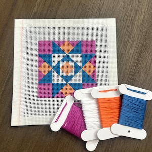May include: A needlepoint canvas with a geometric star pattern in shades of blue, orange, and purple. Spools of thread in matching colors are arranged nearby, including white, orange, blue, and purple. The canvas is on a wooden surface.