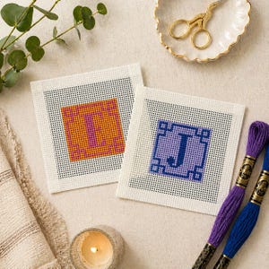 May include: Two square needlepoint canvases with the letters "I" and "J" stitched in orange, pink, and purple thread. The canvases are on a white background, with spools of blue and purple embroidery floss.