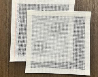 May include: Two white needlepoint canvases with a grid of small holes, one slightly overlapping the other. The edges are finished with a white border. One canvas has a pink stripe along the edge. The background is a dark wood grain.