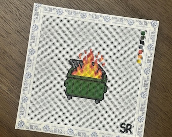 May include: A square needlepoint canvas featuring a pixelated green dumpster on fire with orange and yellow flames. The border is taped with white fabric tape printed with the text SOUTHERN MADE. A color palette key is on the right and SR is in the corner.