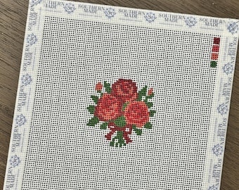 May include: A square needlepoint canvas featuring a central bouquet of three red roses with green leaves and a red ribbon. The white mesh canvas has a border printed with the text Southern Made and a small color palette of red and green squares.