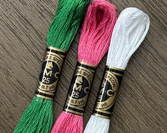 May include: Three skeins of embroidery floss in vibrant green, bright pink, and white. Each skein features a black and gold paper label with the text DMC, Moulin Special, 25, and 8.7 yd. The threads have a smooth, lustrous texture and are arranged side by side.