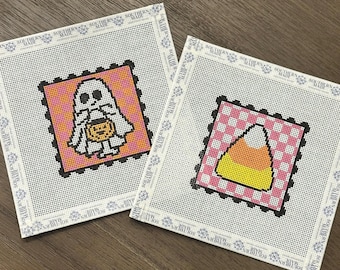 May include: Two square needlepoint canvases with white borders labeled Southern Made. One canvas features a white ghost holding an orange pumpkin on a pink and orange checkered background. The other shows a yellow and orange candy corn on a pink checkered background.
