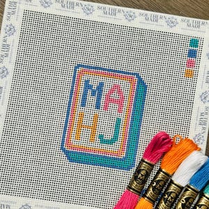 May include: A needlepoint canvas with the letters "MA HJ" in a colorful, outlined design. The canvas is white with a grid pattern, and several skeins of embroidery floss in various colors are arranged below.