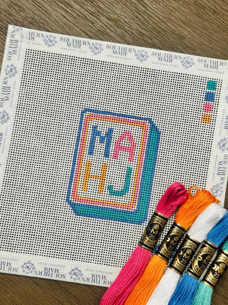 May include: A needlepoint canvas with the letters "MA HJ" in a colorful, outlined design. The canvas is white with a grid pattern, and several skeins of embroidery floss in various colors are arranged below.