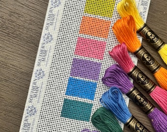 May include: A rectangular needlepoint canvas featuring a vertical column of six colored blocks in yellow, orange, pink, purple, blue, and teal, with the letters SR at the bottom. Six matching embroidery floss skeins are arranged alongside the canvas.