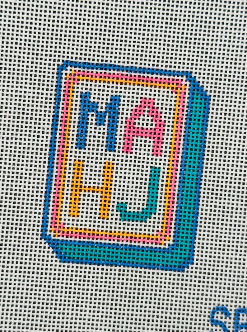 May include: Needlepoint canvas with the letters "MA HJ" in blue, pink, and yellow, framed in a teal border. The letters are stitched on a white background, creating a colorful, geometric design. The canvas is likely for a craft project.