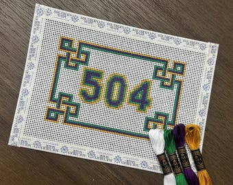 May include: A rectangular needlepoint canvas featuring the number 504 in purple, green, and gold, surrounded by a geometric border. The canvas edges are bound with white tape printed with the text Southern Made. Four skeins of embroidery floss.