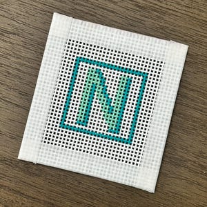 May include: A square needlepoint canvas featuring the letter "N" in teal and light green, framed by a teal border. The background is a grid of black and white squares, with a white border around the edge.