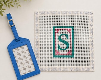 May include: A bright blue luggage tag with a gold-toned buckle and a square needlepoint canvas featuring a central letter S in dark green, surrounded by a pink border and a teal frame, with the text SOUTHERN MADE repeated along the edges.
