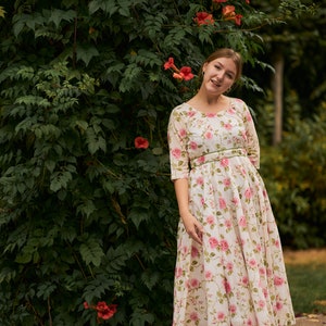 May include: A woman wearing a white floral dress with a green belt stands in front of a green vine covered wall with pink flowers.