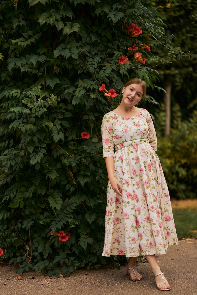 May include: A woman wearing a white floral dress with a green belt stands in front of a green vine covered wall with pink flowers.