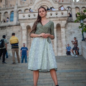 May include: A woman wearing a green and white patterned dress stands on a set of stone steps in front of a large building. She is looking directly at the camera.
