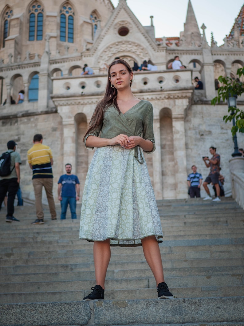 May include: A woman wearing a green and white patterned dress stands on a set of stone steps in front of a large building. She is looking directly at the camera.