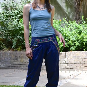 May include: A woman wearing a light blue tank top and dark blue pants with a colorful embroidered waistband. She is standing in a grassy area with a brick wall and greenery in the background.