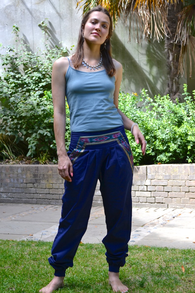 May include: A woman wearing a light blue tank top and dark blue pants with a colorful embroidered waistband. She is standing in a grassy area with a brick wall and greenery in the background.