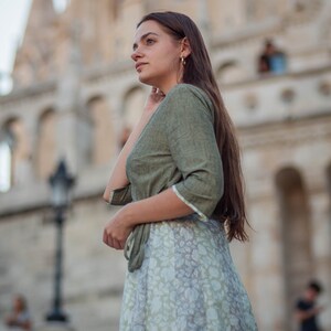 May include: A woman wearing a green top and a floral skirt stands in front of a large building. She is looking to the right of the frame.