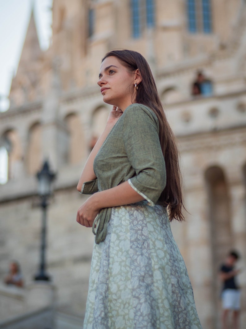 May include: A woman wearing a green top and a floral skirt stands in front of a large building. She is looking to the right of the frame.