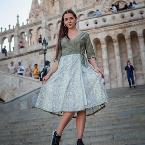 May include: A woman wearing a green and white floral wrap dress with a white underskirt, standing on a set of stone steps in front of a large stone building.