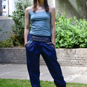 May include: A woman wearing a blue tank top and dark blue pants with a colorful embroidered waistband. She is standing in a grassy area with a brick wall in the background.
