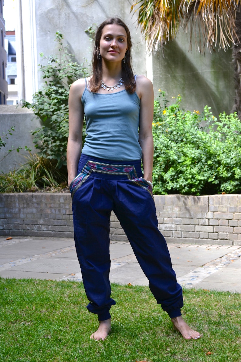 May include: A woman wearing a blue tank top and dark blue pants with a colorful embroidered waistband. She is standing in a grassy area with a brick wall in the background.