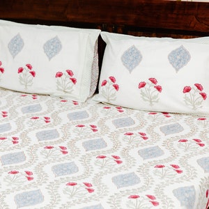 May include: White bed linen set with a floral pattern, including two pillowcases. The pillowcases display red flowers and blue leaf designs. The bedsheet has a repeating pattern of the same elements. The linens are on a dark wooden bed frame.