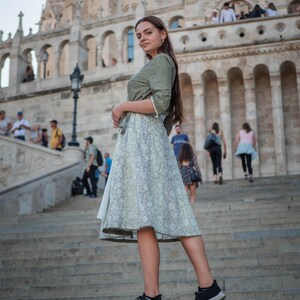 May include: A woman wearing a green and white floral dress and black trainers stands on a set of stone steps in front of a large stone building.