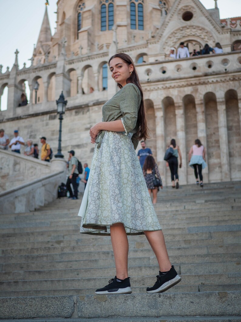 May include: A woman wearing a green and white floral dress and black trainers stands on a set of stone steps in front of a large stone building.