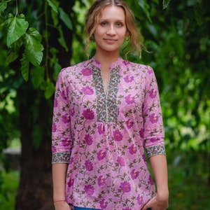 May include: A light pink tunic top with a floral pattern in shades of purple and green. The top has a mandarin collar and three-quarter sleeves. The neckline and sleeve cuffs are trimmed with a patterned border. The model is wearing blue jeans.