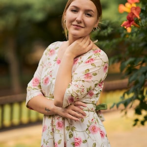May include: A woman wearing a white dress with a floral pattern, standing in front of a green bush with orange flowers. She has her hand on her chin and is looking at the camera. The dress has a green sash tied at the waist. The woman has a tattoo on her arm that says "hope".