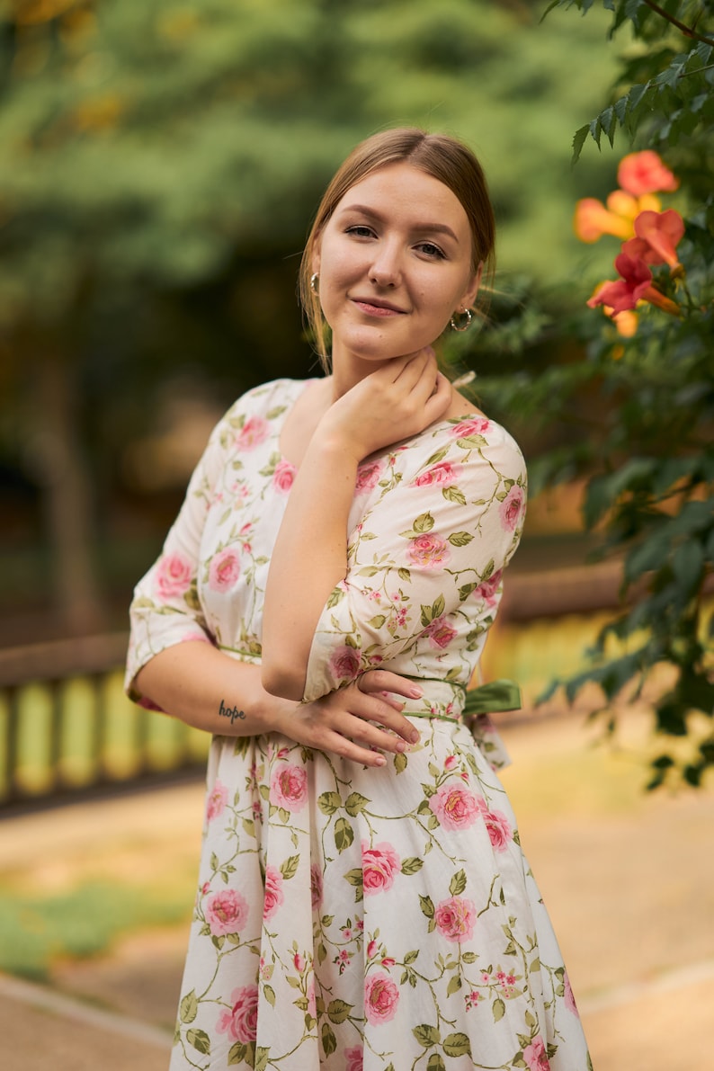 May include: A woman wearing a white dress with a floral pattern, standing in front of a green bush with orange flowers. She has her hand on her chin and is looking at the camera. The dress has a green sash tied at the waist. The woman has a tattoo on her arm that says "hope".