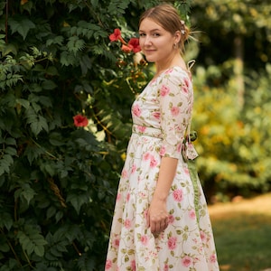 May include: A woman wearing a white dress with a floral pattern and a green sash. The dress has a scoop neckline and 3/4 sleeves. She is standing in front of a green leafy background with pink flowers.
