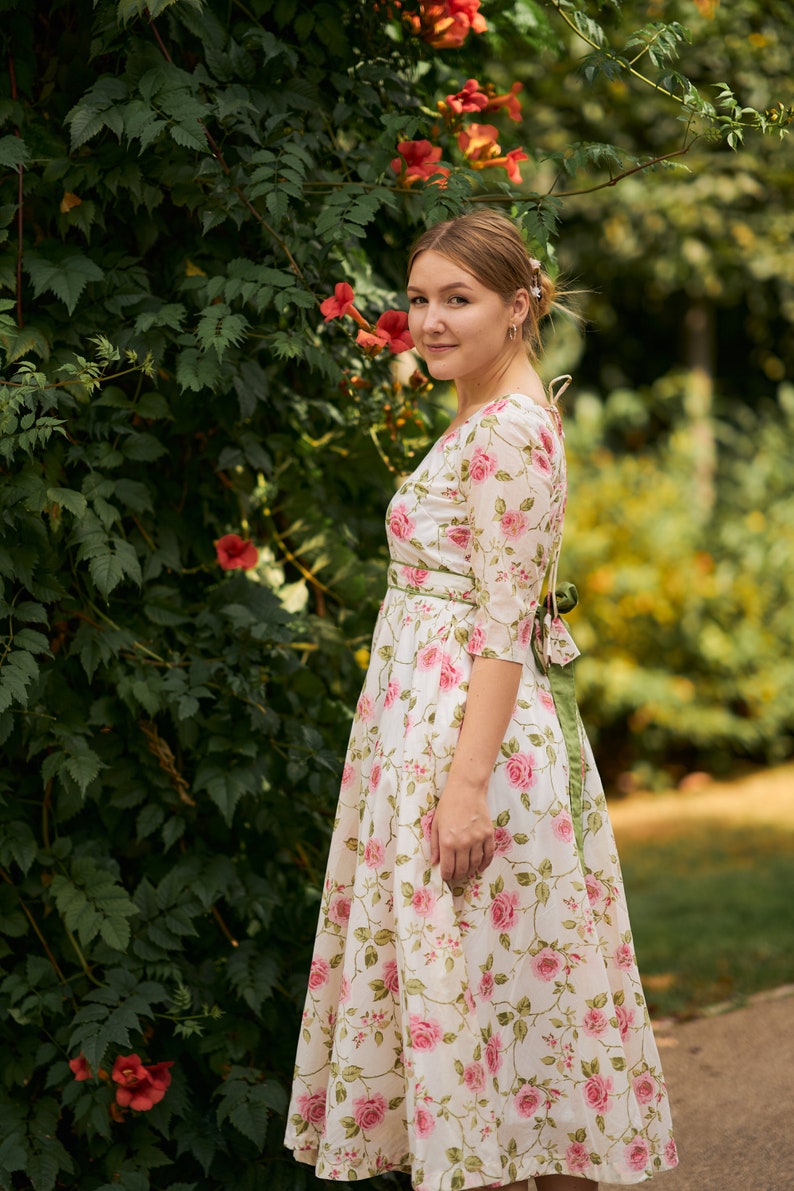 May include: A woman wearing a white dress with a floral pattern and a green sash. The dress has a scoop neckline and 3/4 sleeves. She is standing in front of a green leafy background with pink flowers.