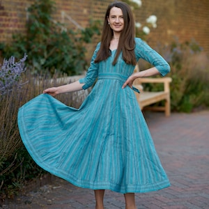 May include: A woman wearing a blue and white striped dress with a cinched waist and a flowing skirt. She is standing on a brick pathway with a brick wall and greenery in the background.
