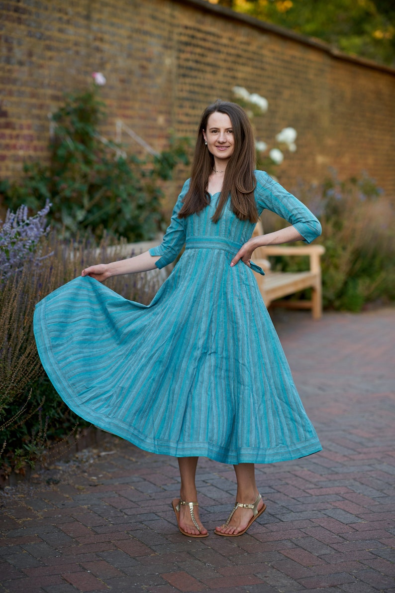 May include: A woman wearing a blue and white striped dress with a cinched waist and a flowing skirt. She is standing on a brick pathway with a brick wall and greenery in the background.