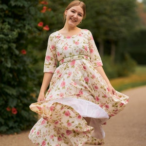 May include: A woman in a white dress with pink floral print and a green sash is twirling in a park. The dress has a full skirt and is flowing in the wind.