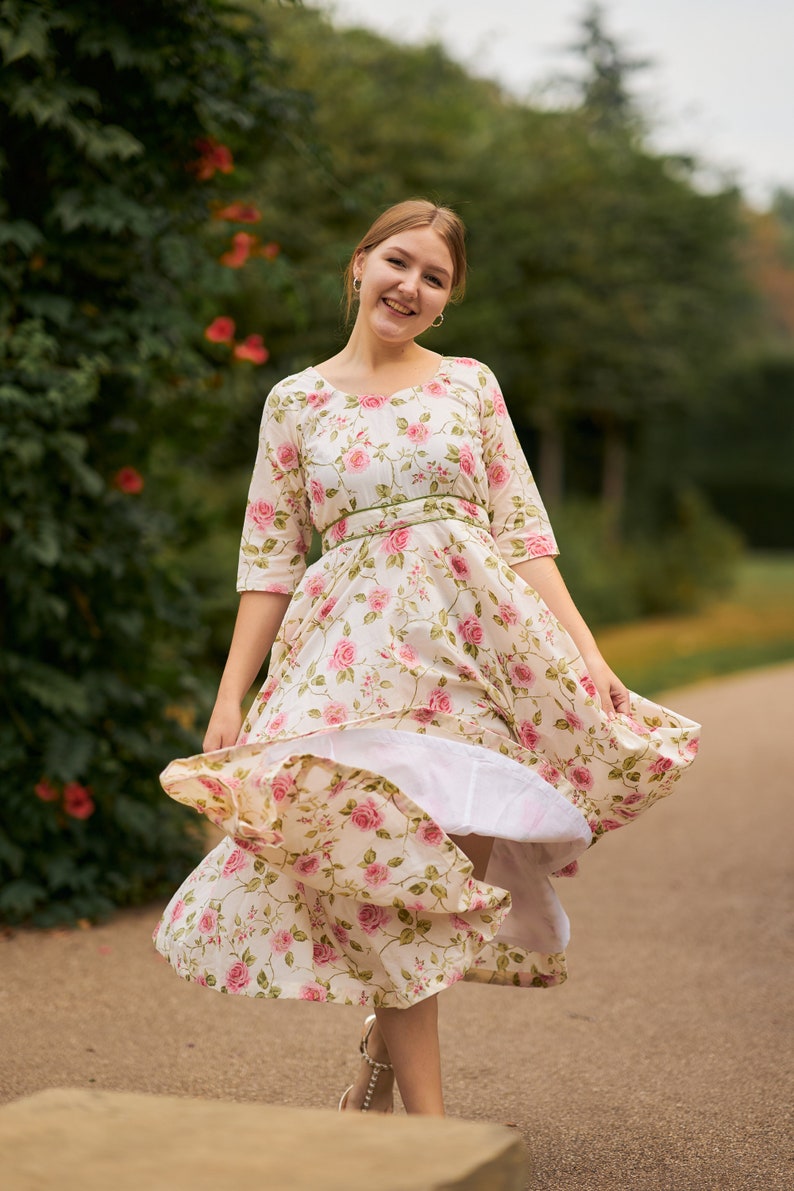 May include: A woman in a white dress with pink floral print and a green sash is twirling in a park. The dress has a full skirt and is flowing in the wind.
