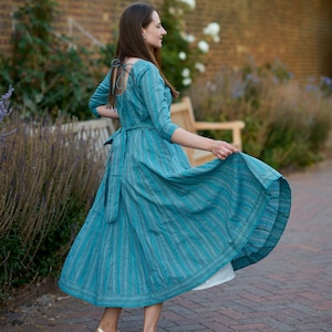 May include: A woman wearing a teal blue dress with white stripes, standing on a brick pathway. The dress has a low back and a tie at the waist. She is wearing gold sandals.