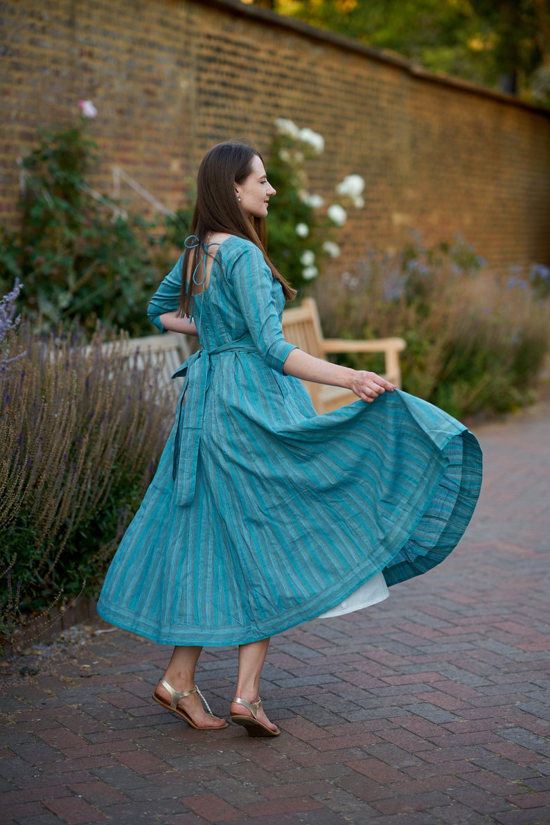 May include: A woman wearing a teal blue dress with white stripes, standing on a brick pathway. The dress has a low back and a tie at the waist. She is wearing gold sandals.
