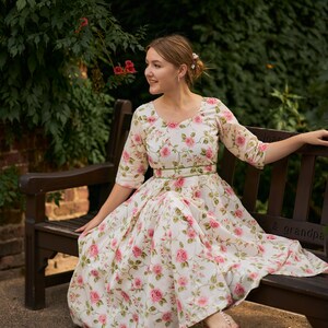 May include: A woman wearing a white dress with pink floral print sits on a wooden bench. The dress has a green sash around the waist and the woman is wearing gold sandals.