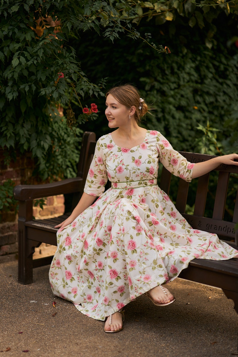 May include: A woman wearing a white dress with pink floral print sits on a wooden bench. The dress has a green sash around the waist and the woman is wearing gold sandals.