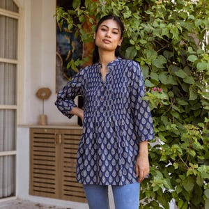 Peut inclure: Une femme portant une tunique bleu et blanc à motifs avec une fermeture boutonnée et des manches trois quarts. Elle porte également un jean bleu clair.