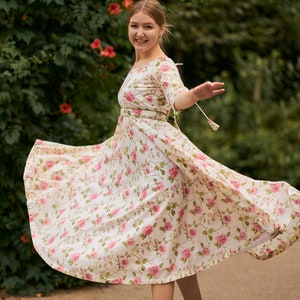 May include: A woman wearing a white dress with a pink floral pattern and gold sandals. The dress has a cinched waist and a flowing skirt. She is standing in front of a green leafy background.