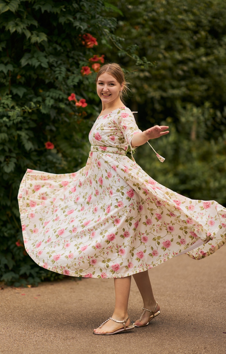 May include: A woman wearing a white dress with a pink floral pattern and gold sandals. The dress has a cinched waist and a flowing skirt. She is standing in front of a green leafy background.